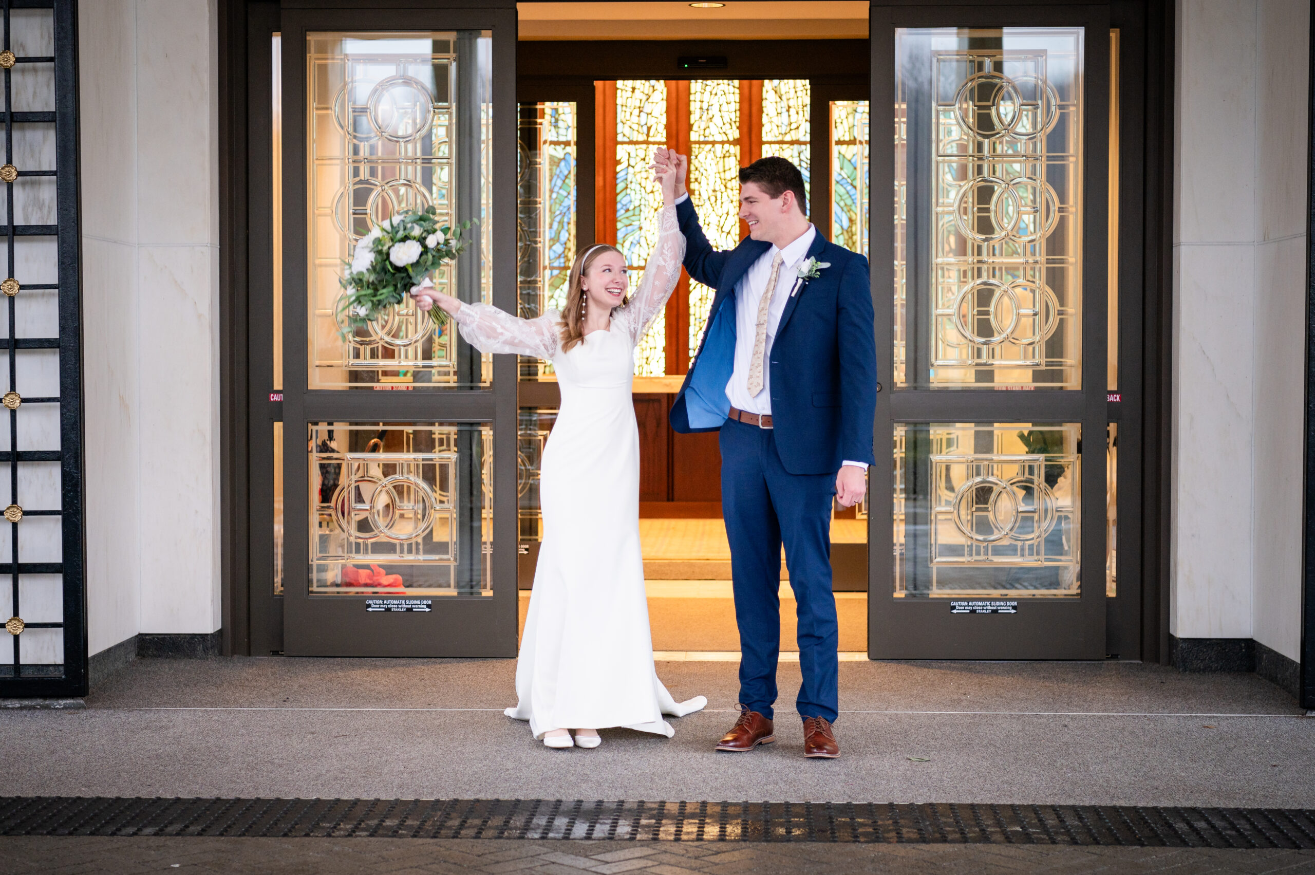 Bride In White Dress And Groom In Blue Suit Stand Outside Building
