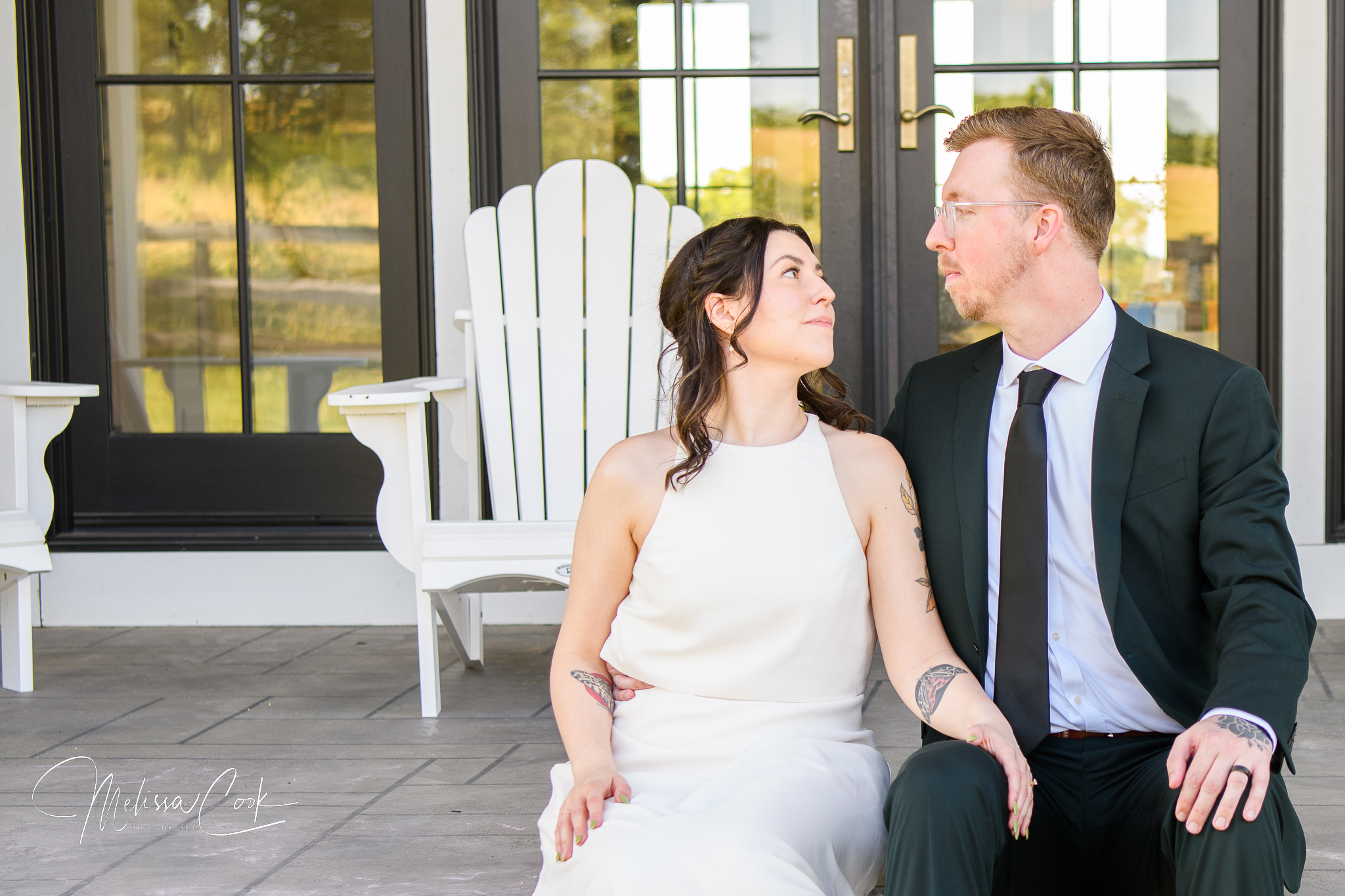 A couple in wedding attire sits on a porch in front of white chairs and glass doors, gazing at each other lovingly. Capturing this special moment, Melissa Cook Weddings highlights the elegance of her white dress and his dark suit and glasses.