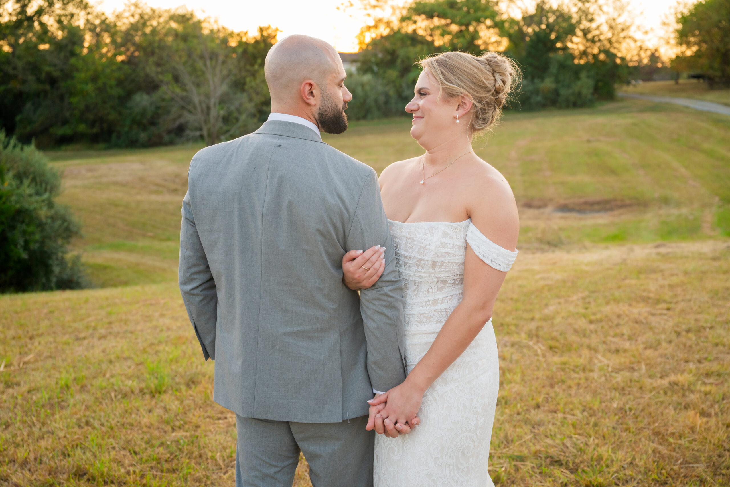 A bride in a white off-shoulder gown smiles at her groom in a gray suit as they hold hands in a sunlit field at golden hour, surrounded by trees—a magical moment captured by Melissa Cook Weddings.
