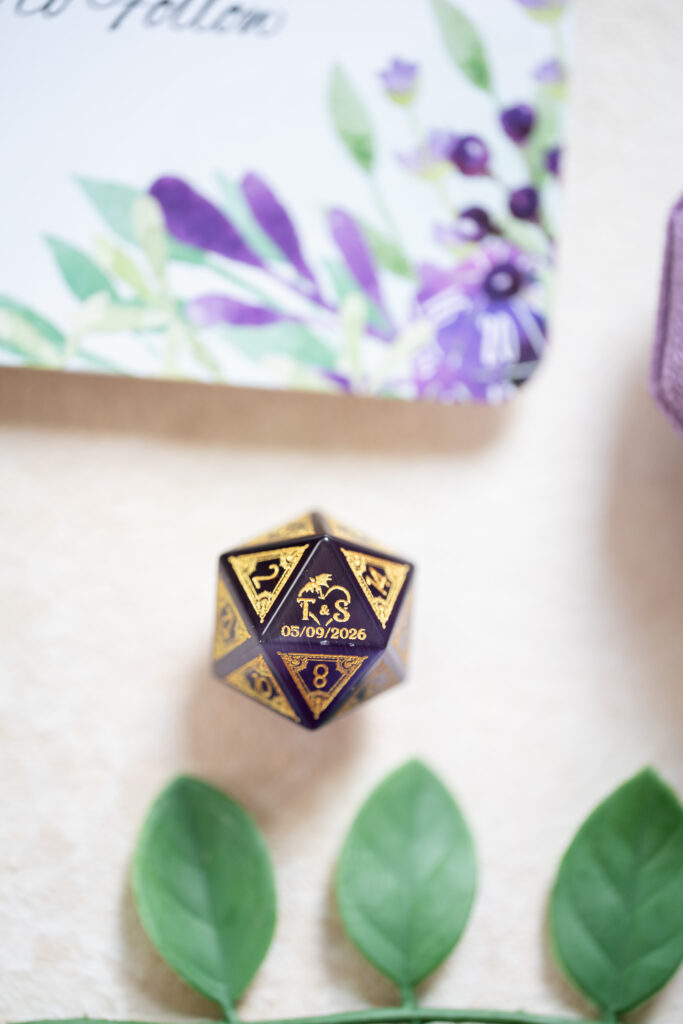 A black and gold 20-sided die with a dragon design and the date 05/09/2026 is placed on a light surface, surrounded by green leaves and a floral-patterned card.