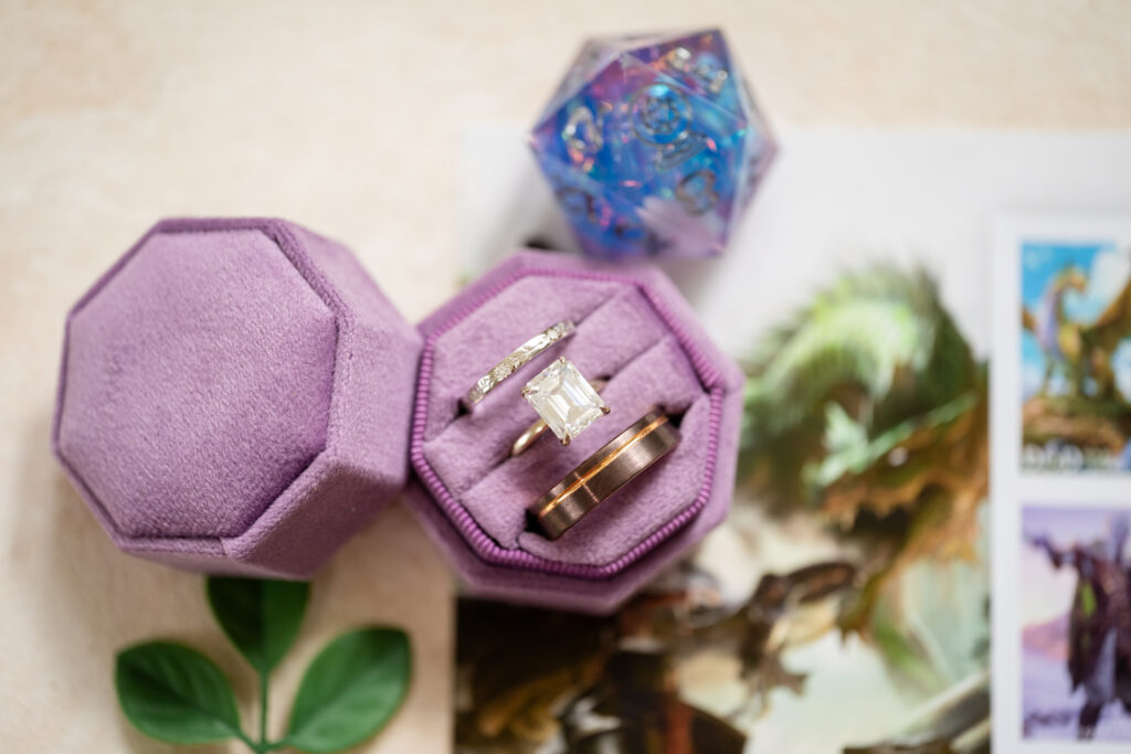 A pair of rings, one with a large rectangular gemstone and one plain, are displayed in a purple hexagonal ring box. Nearby are a green leaf, a colorful dice, and fantasy-themed illustrations.