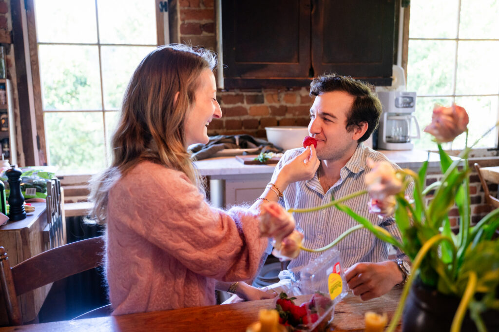 A woman playfully feeds a strawberry to a smiling man at a wooden kitchen table, with flowers and kitchen items visible in the background. They appear to be enjoying a lighthearted moment together.