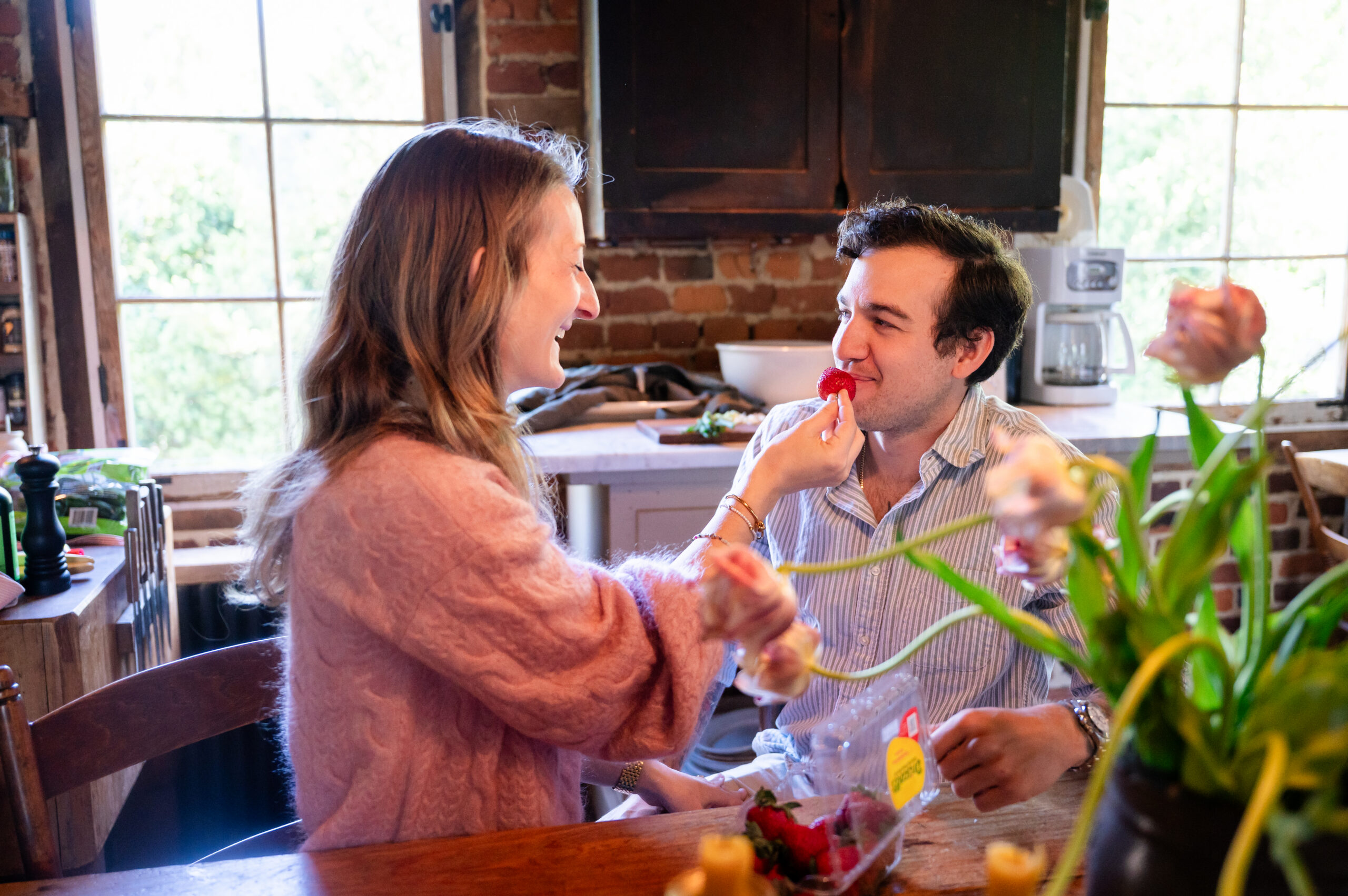 A woman playfully feeds a strawberry to a smiling man at a wooden kitchen table, with flowers and kitchen items visible in the background. They appear to be enjoying a lighthearted moment together.