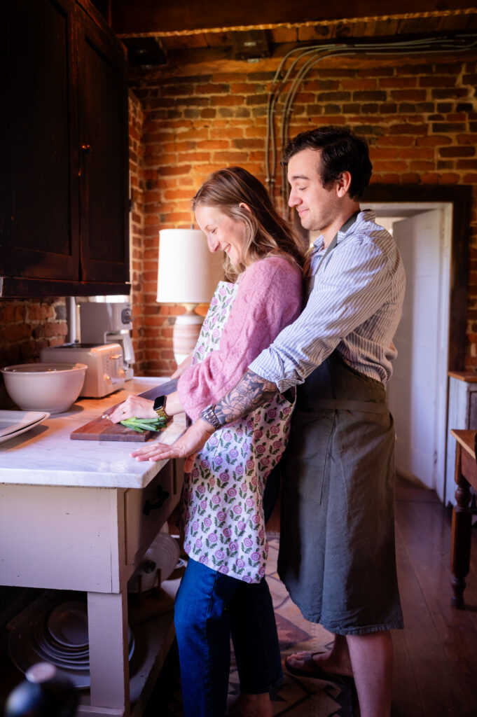 A man stands behind a smiling woman as she chops vegetables on a kitchen counter. Both wear aprons and appear to be enjoying cooking together in a cozy, rustic kitchen with exposed brick walls.