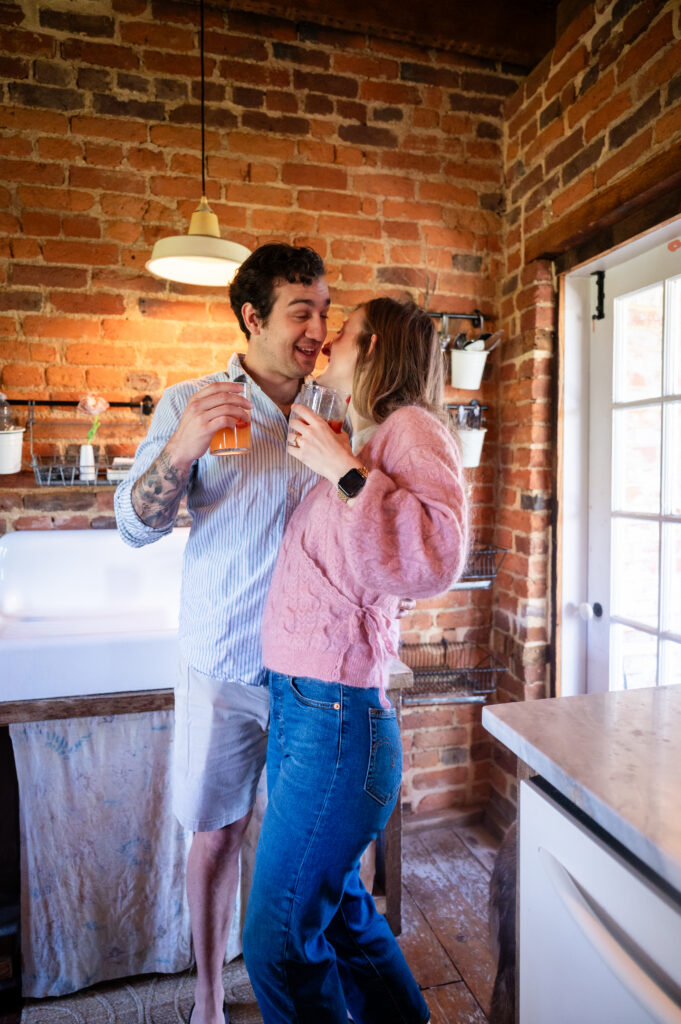 A smiling couple stands in a rustic brick kitchen, holding drinks and leaning in close to each other, appearing happy and affectionate. Natural light streams in through a nearby door.