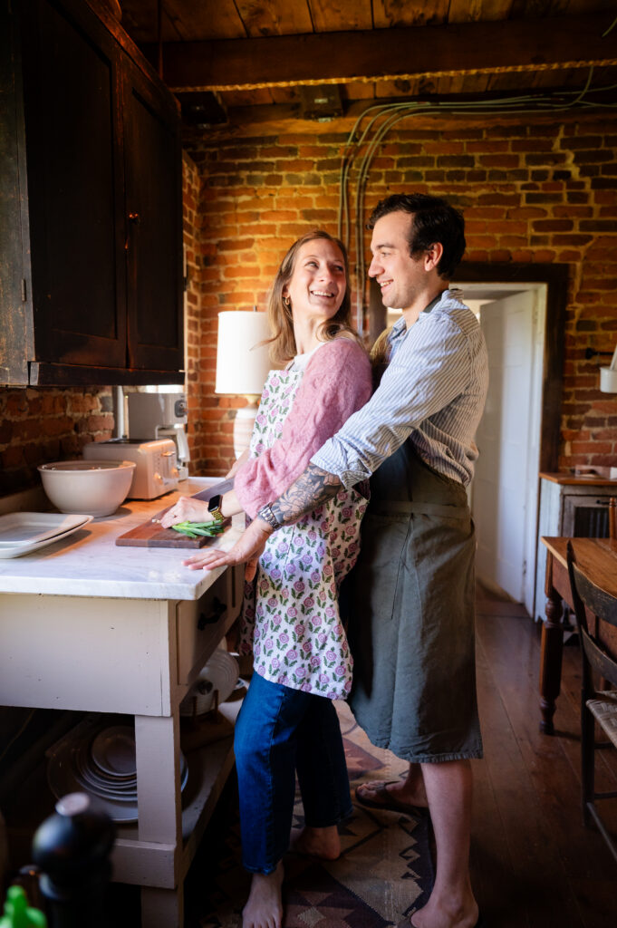 A smiling couple stands together in a rustic kitchen with exposed brick walls. The woman chops vegetables on a counter while the man, wearing an apron, embraces her from behind. Both look happy and relaxed.