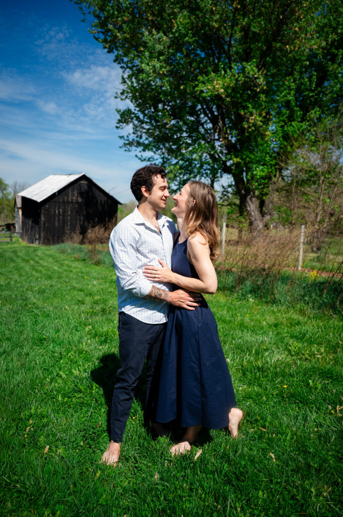 A couple stands barefoot on green grass, smiling and embracing each other on a sunny day. A rustic wooden barn and tall trees are in the background under a blue sky with scattered clouds.