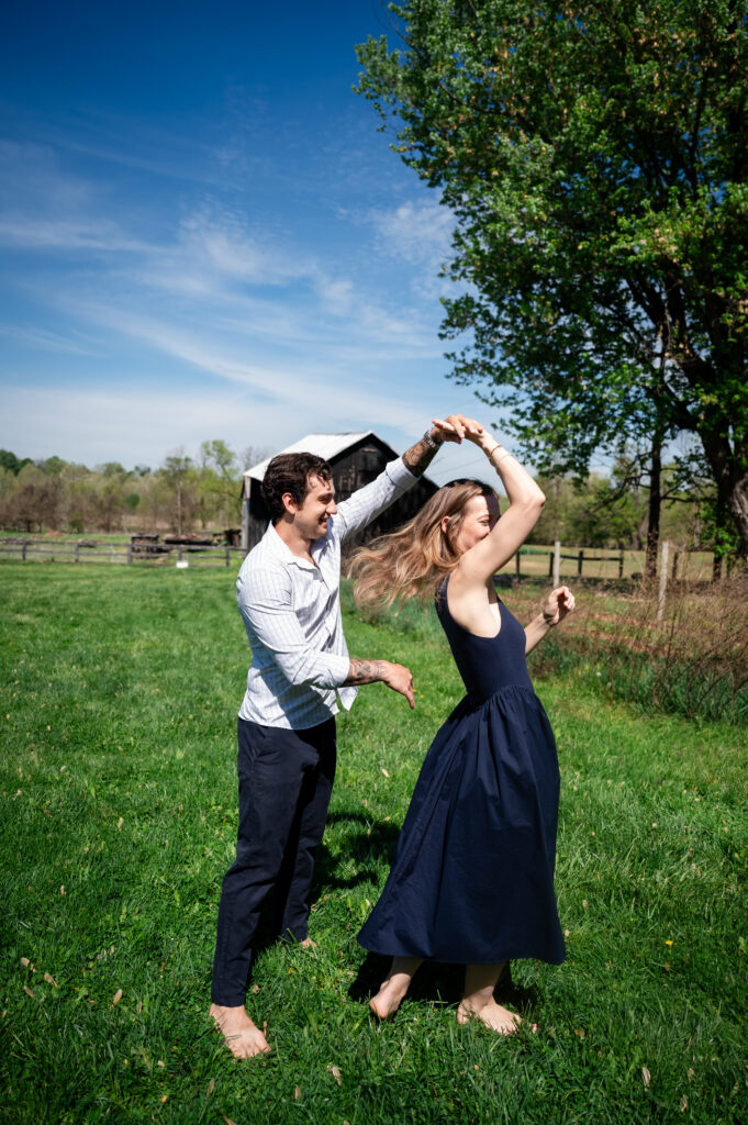A barefoot man and woman dance together on green grass in a sunny, rural field with a wooden barn and trees in the background. The woman twirls, holding the mans hand, both smiling and joyful.