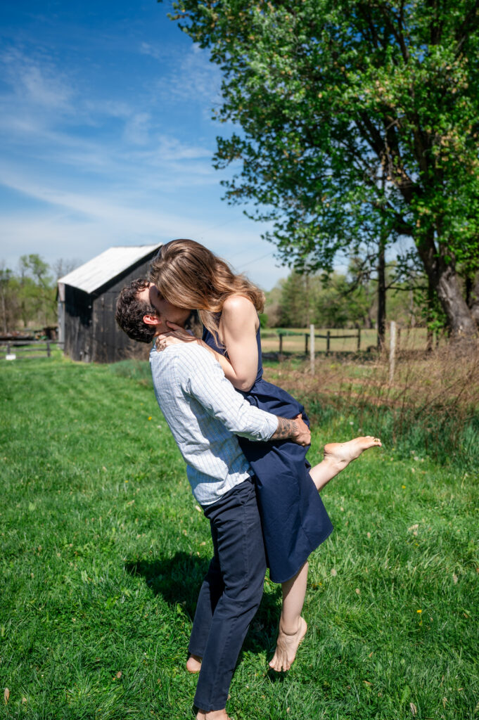A couple embraces and kisses in a grassy field on a sunny day. The man lifts the woman off the ground as she wraps her arms around his neck. A rustic barn and large tree are in the background.